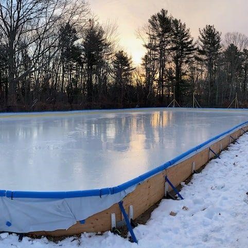 The original back-yard style community rink at sunset, framed by pines and snow.